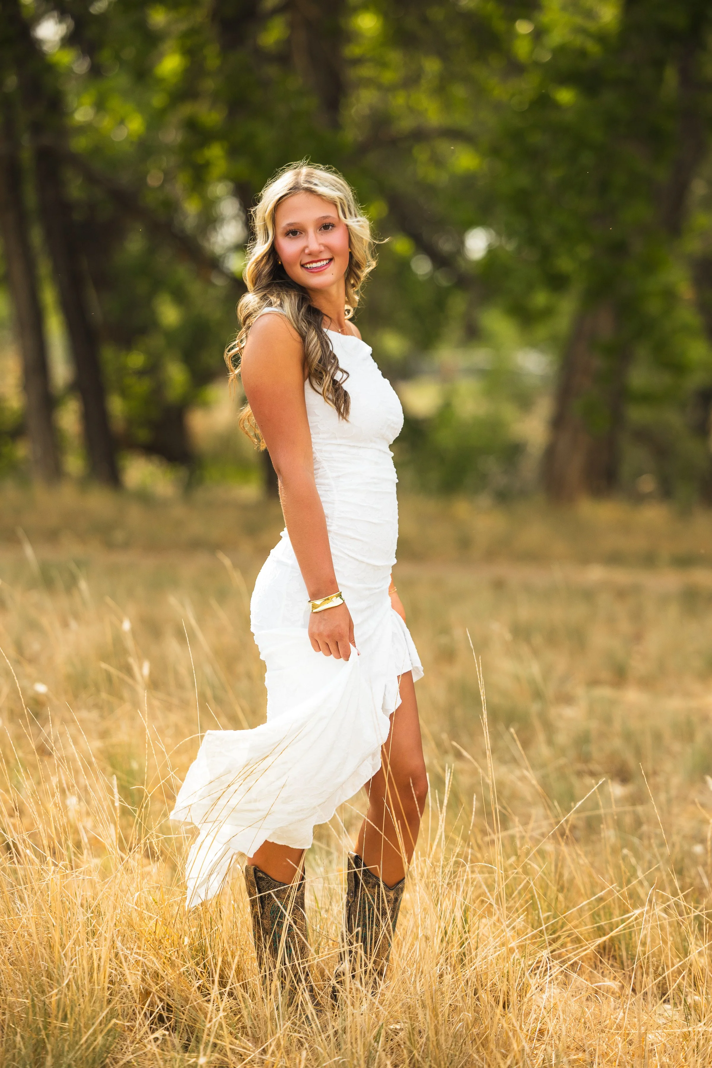 A young woman with long, wavy blonde hair standing in a field of tall, dry grass with a background of green trees, wearing a white dress and cowboy boots, smiling at the camera.