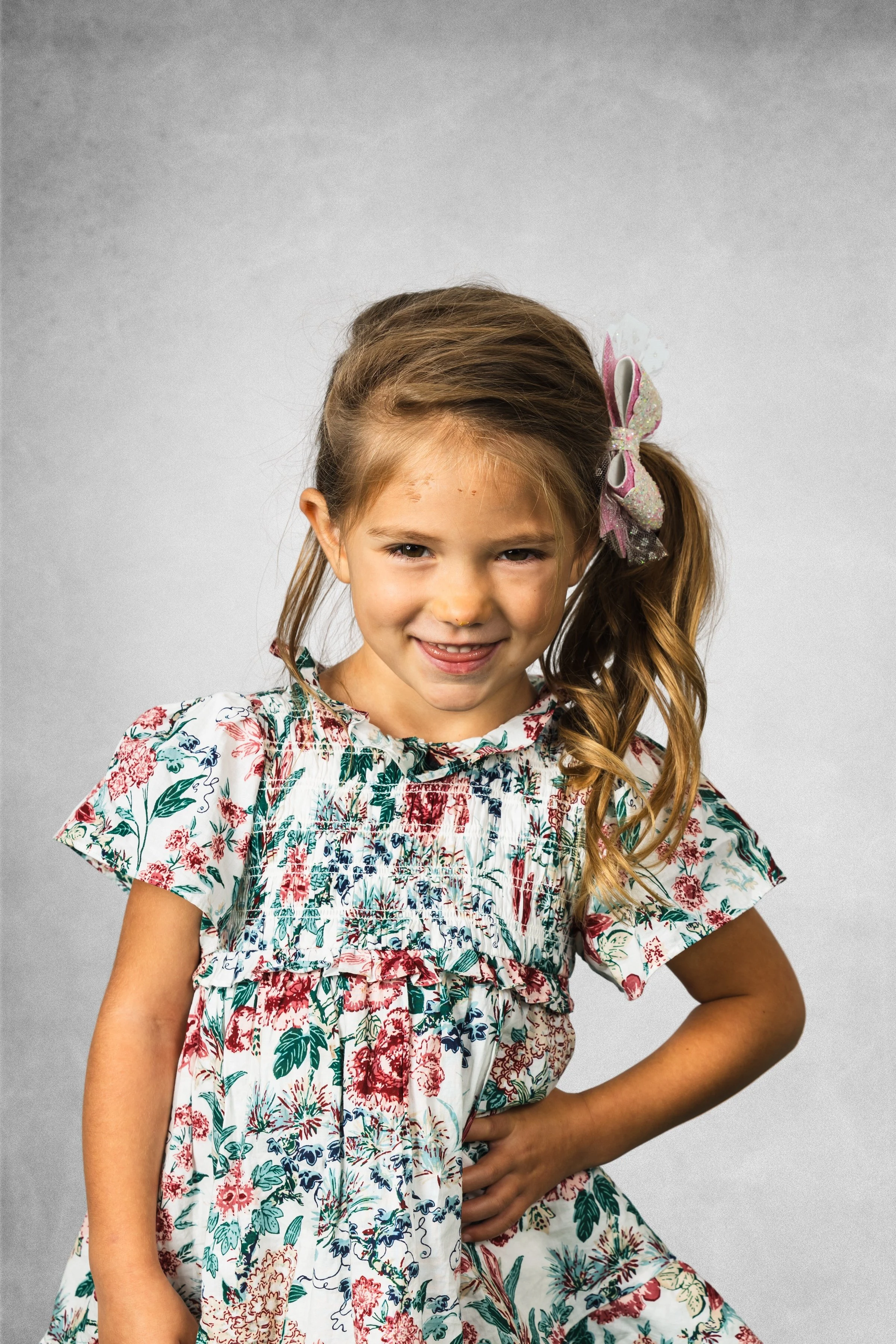 A young girl with long, wavy brown hair in a side ponytail, decorated with a pink and silver glittery butterfly hair accessory. She is wearing a floral dress with a mix of red, green, and blue patterns on a white background. She has a happy expression with a slight smile, one hand on her hip, and is standing against a gray background.