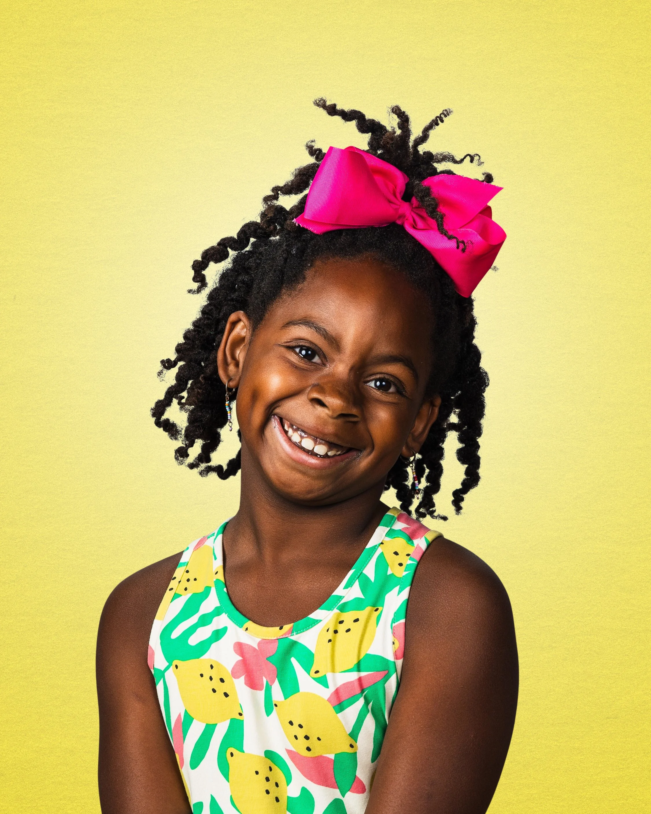A young girl smiling with a missing tooth, wearing a colorful dress with fruit and plant patterns, a bright pink bow in her hair, and colorful earrings, against a yellow background.