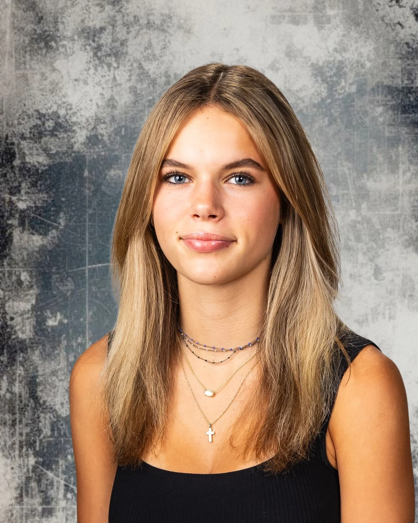 A young woman with shoulder-length blonde hair, blue eyes, and light makeup, wearing a black sleeveless top and layered necklaces, standing against a textured gray background.
