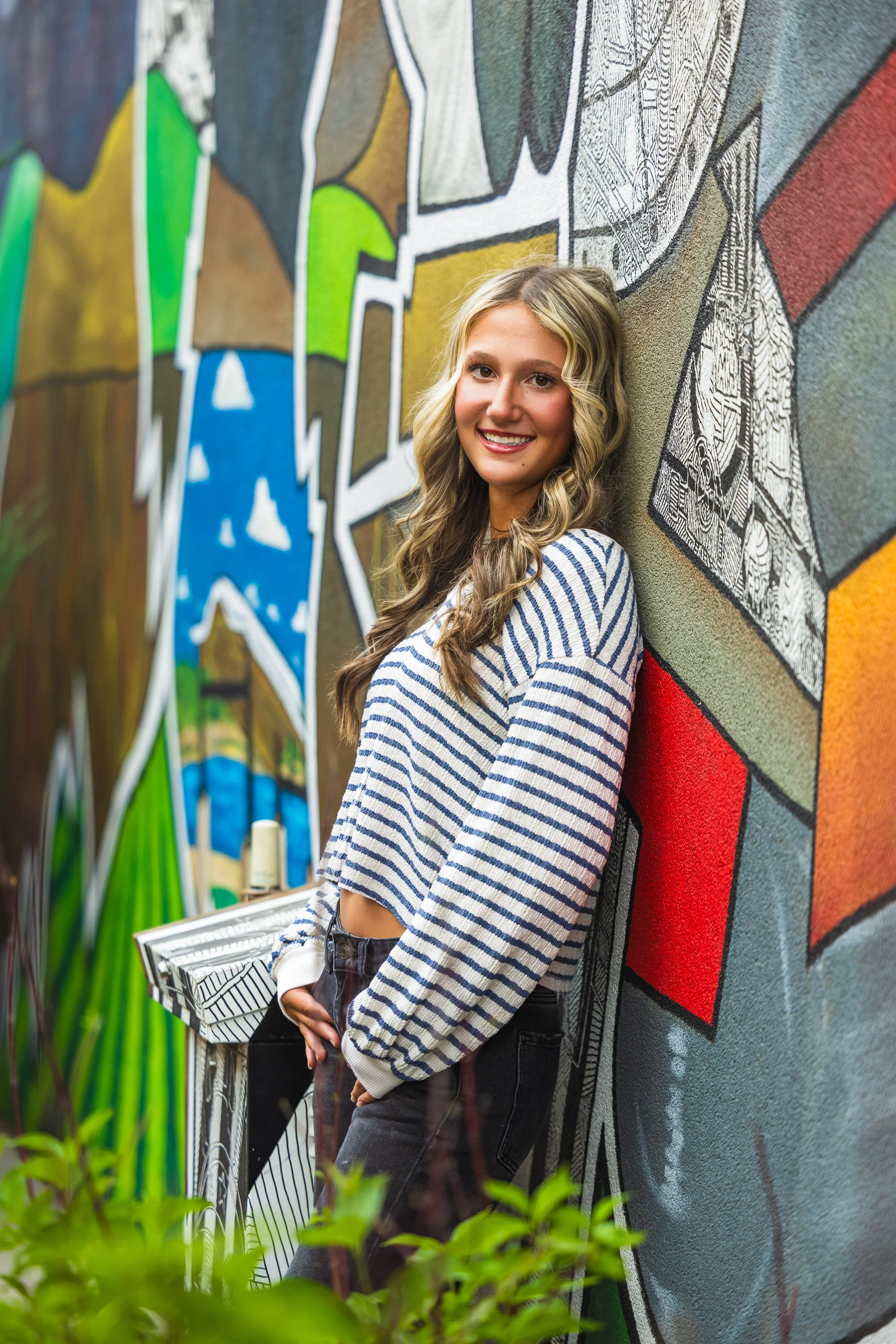 A young woman with long wavy blonde hair, wearing a blue and white striped sweater and black pants, leaning against a colorful graffiti wall and smiling at the camera.