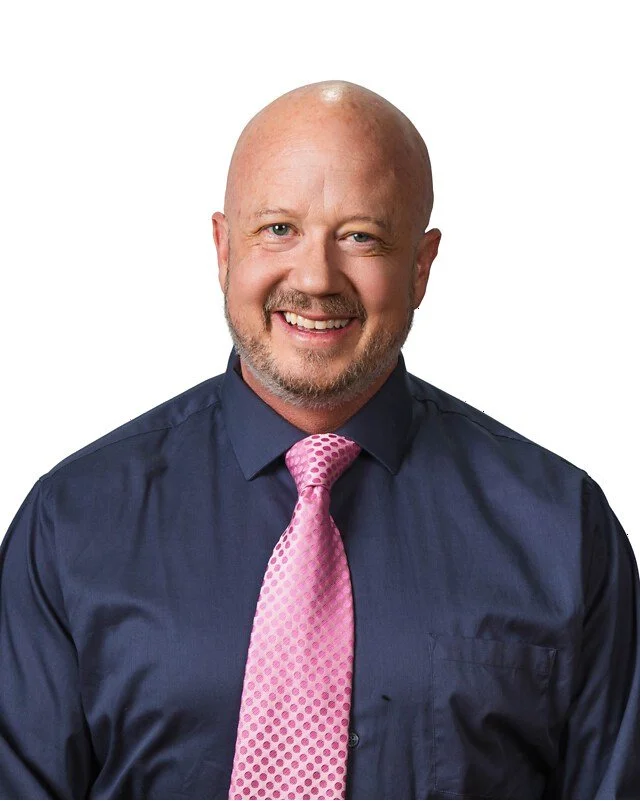 A smiling bald man with a beard, wearing a navy blue dress shirt and a pink polka dot tie, against a white background.