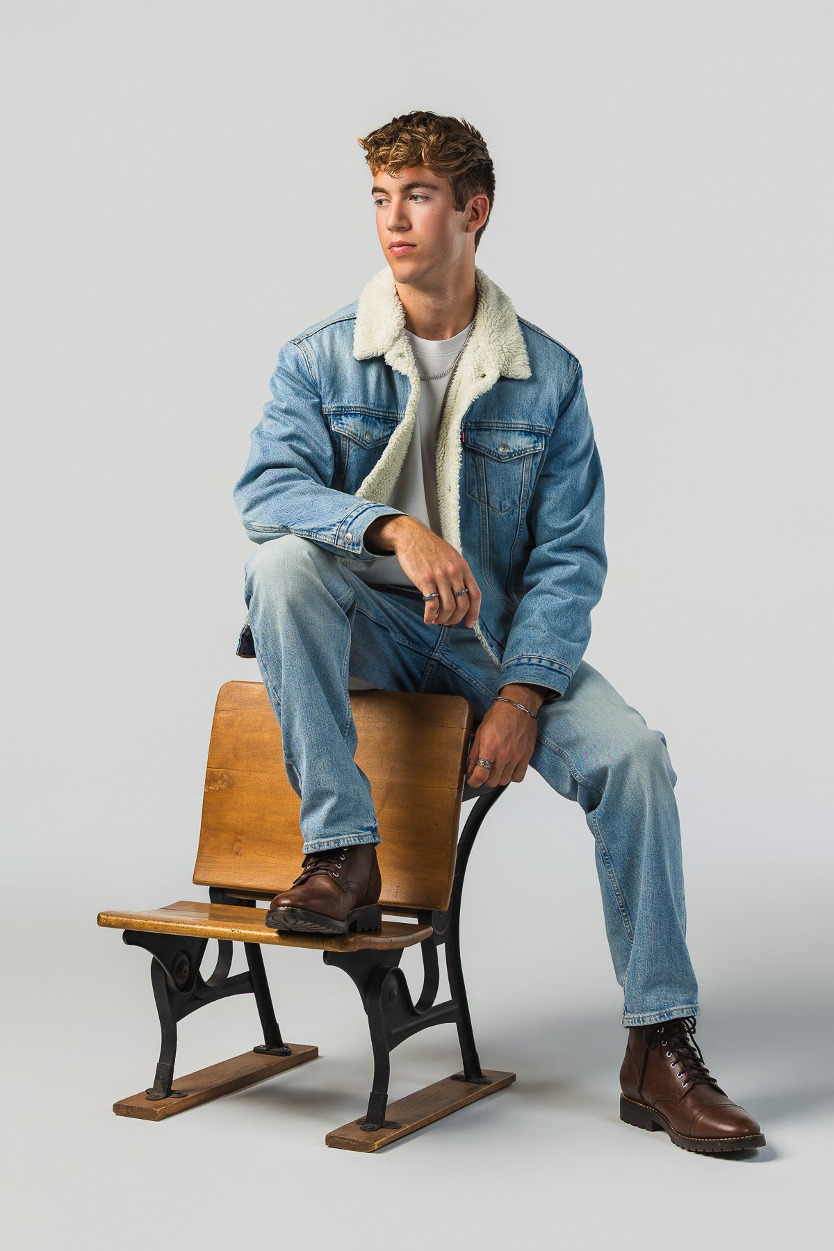 A young man with curly hair sitting on a chair with one foot on the chair's seat. He is wearing a denim jacket with a shearling collar, jeans, and brown boots, against a plain white background.