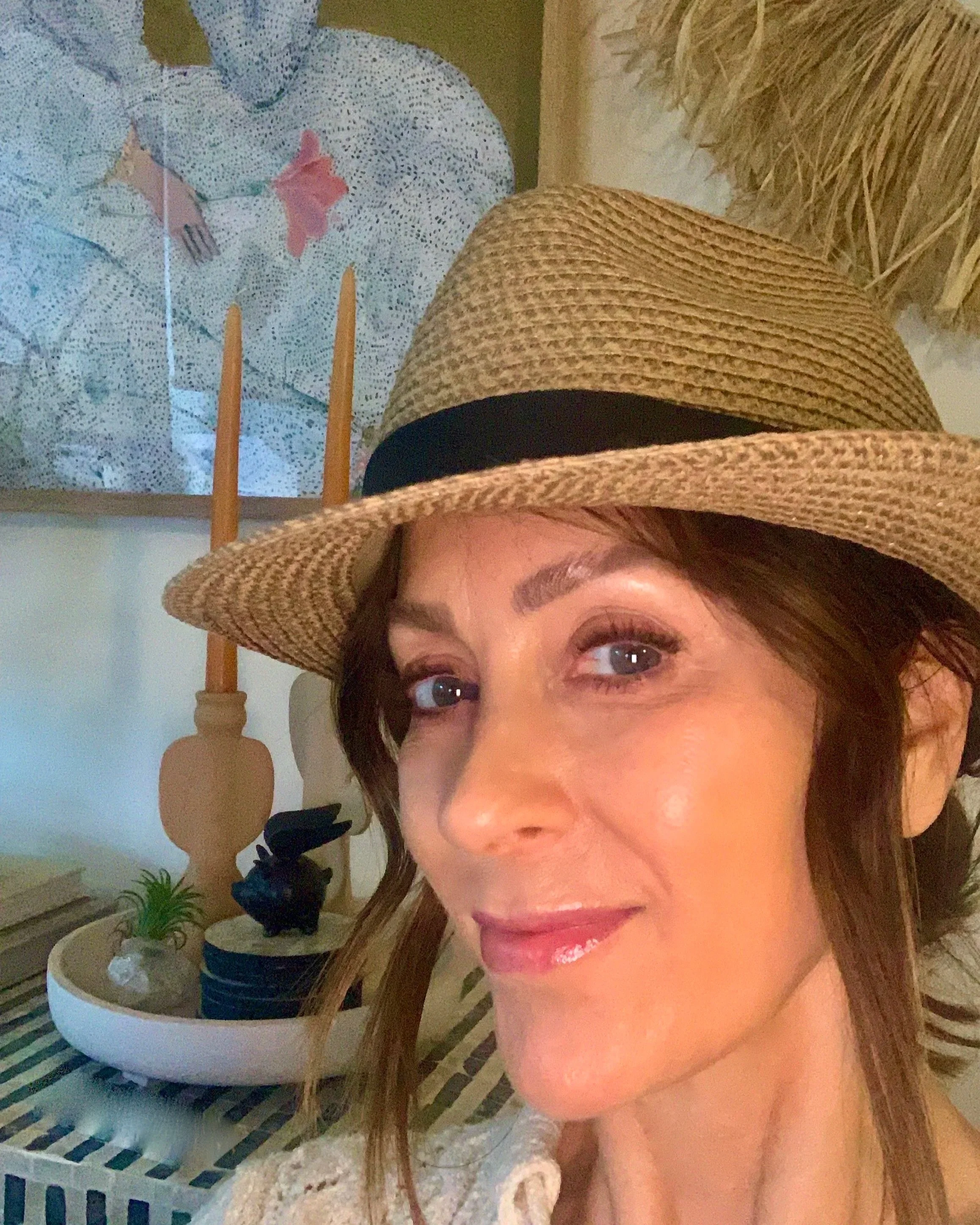 A woman with long brown hair wearing a straw hat smiling, with indoor plants and a decorative lamp in the background.
