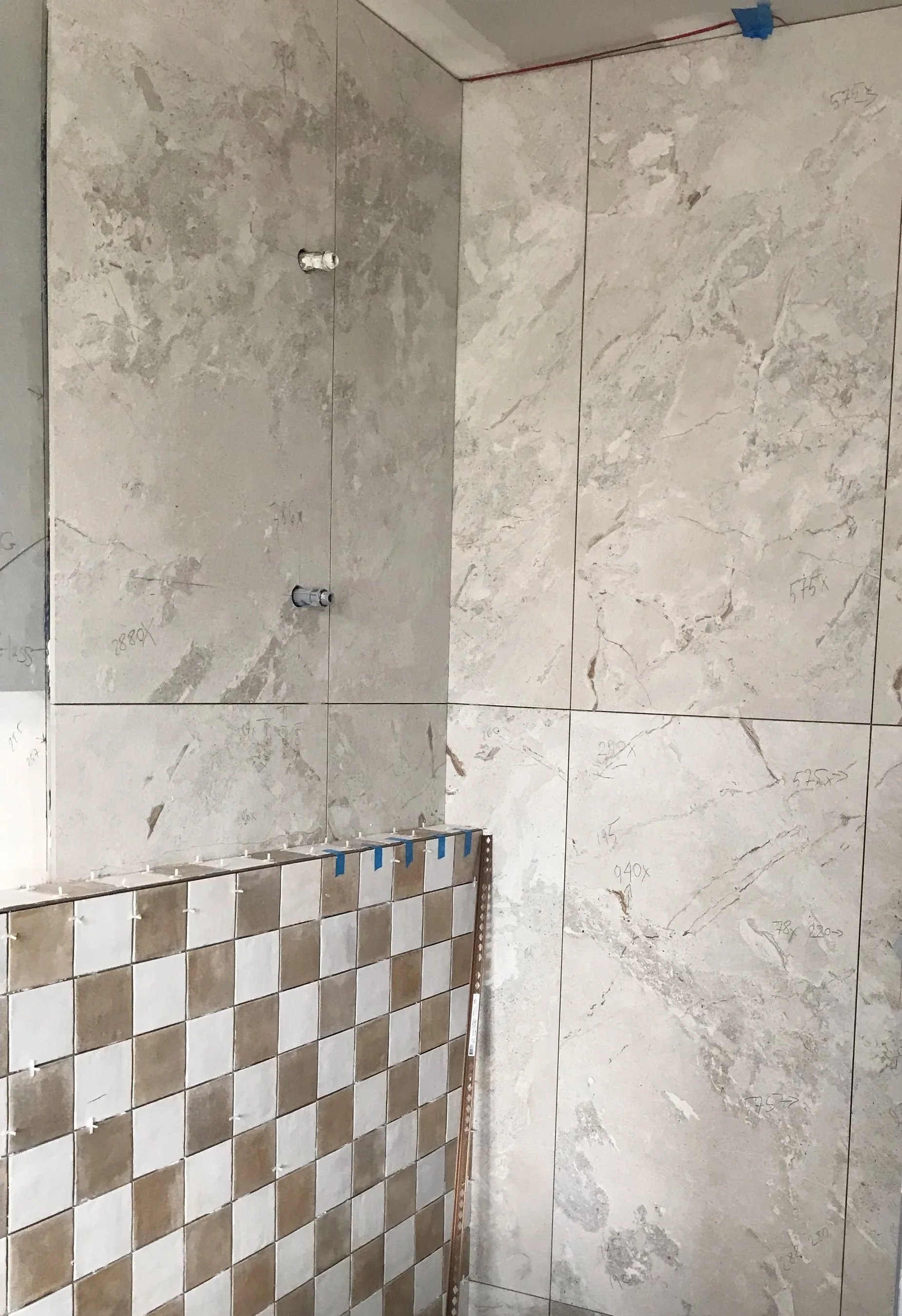 Construction site with large beige marble tiles on the wall, some tiles with handwritten measurements in pencil, and a partial wall with a tiled section in a checkerboard pattern of beige and white. Pipes and construction tools are visible.