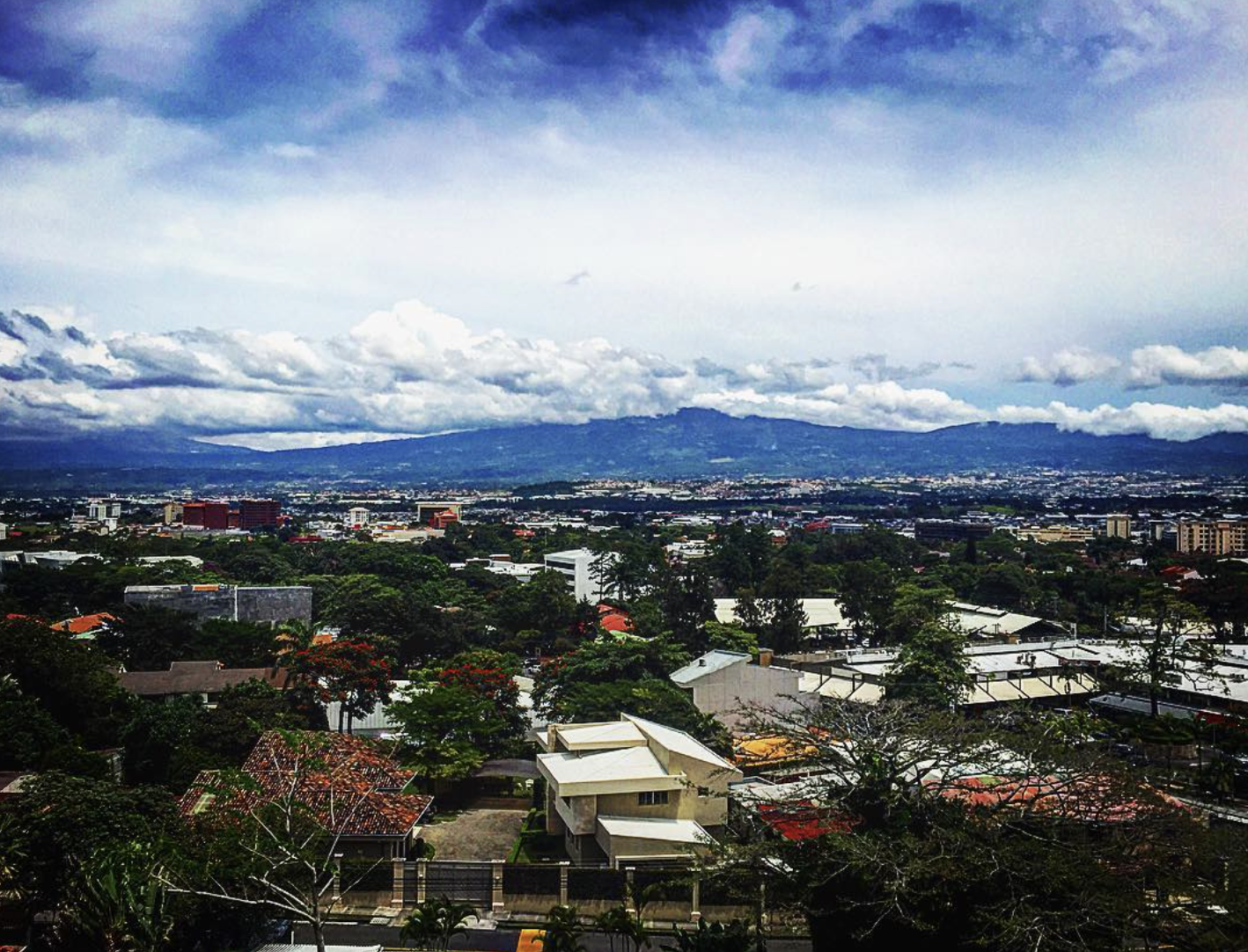View from a hillside terrace in Escazú, Costa Rica, across tree-filled neighbourhoods to mountains under heavy cloud.