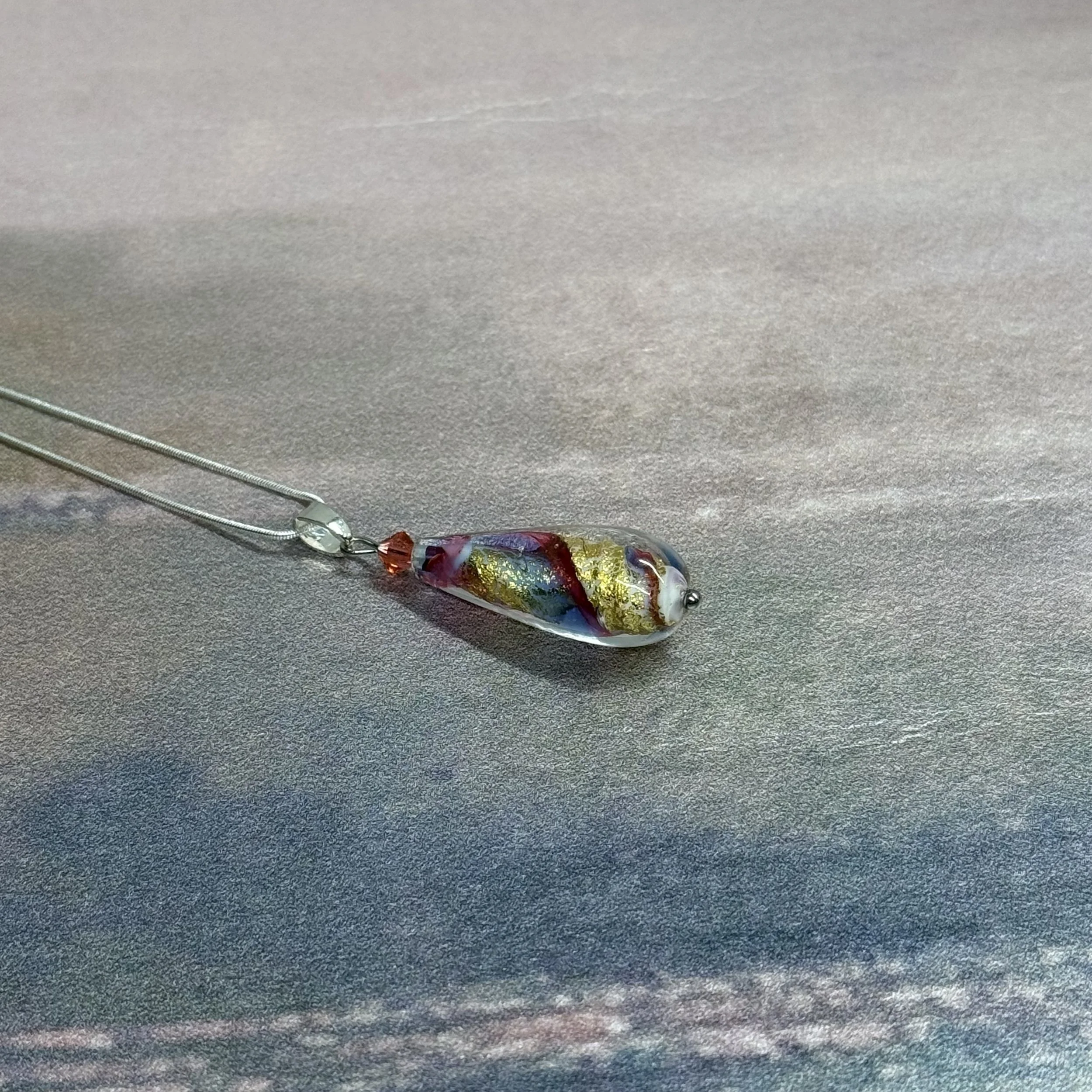 A multi coloured Murano glass teardrop pendant on a sterling silver chain displayed on top of a grey cotton cloth bag with the words Planeterf Jewellery stamped in silver on the bag