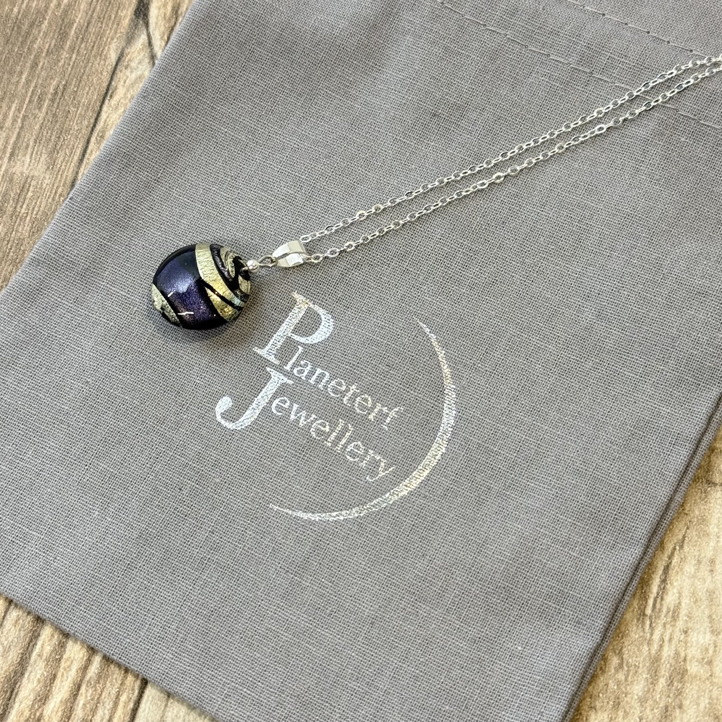 A Murano glass round cushion with purple, black & silver swirls hung on a sterling silver chain. The background is a grey cotton bag stamped with Planeterf Jewellery in silver.