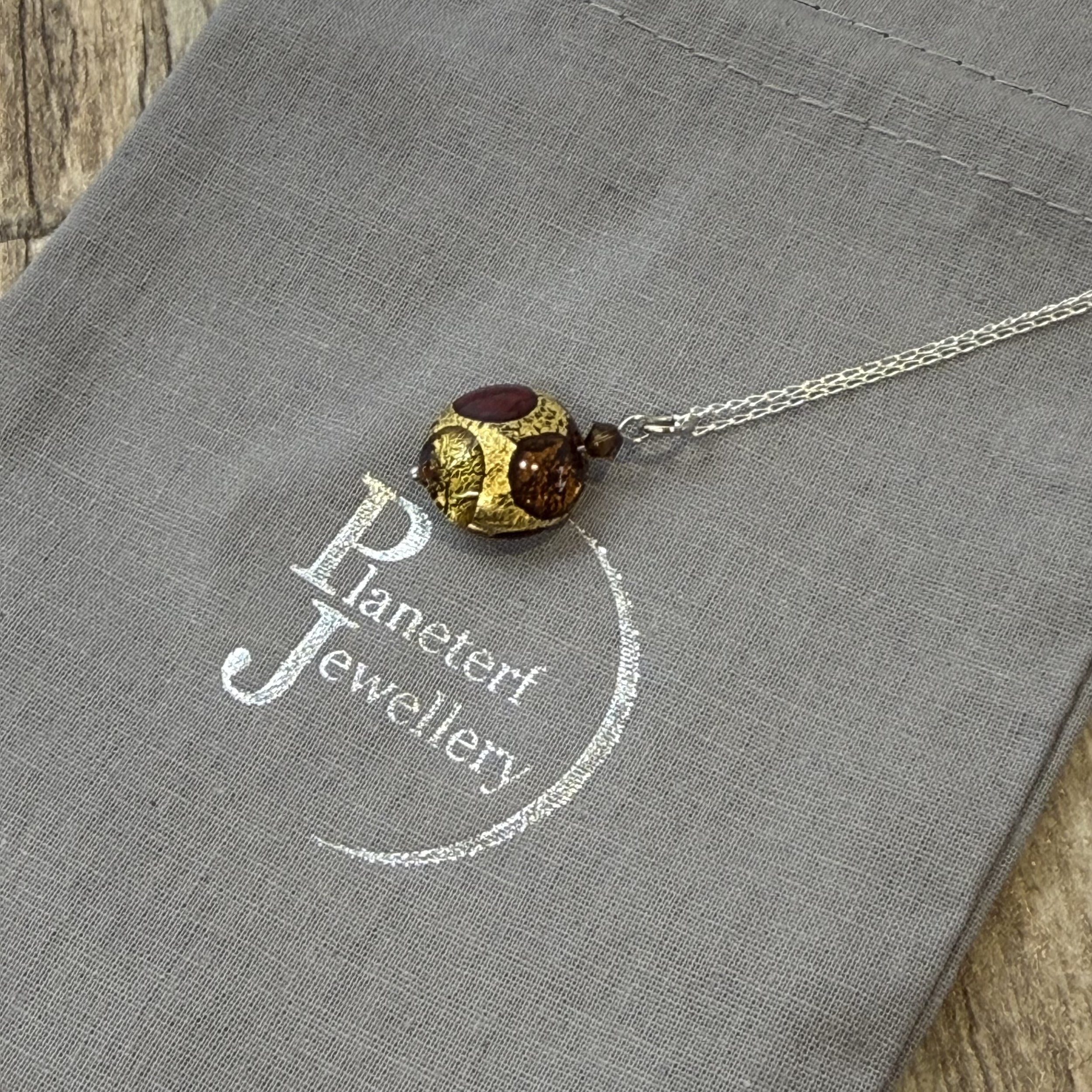 A Murano glass cushion in gold, amber & red colours on a sterling silver chain.hung on a sterling silver chain. The background is a grey cotton bag stamped with Planeterf Jewellery in silver.