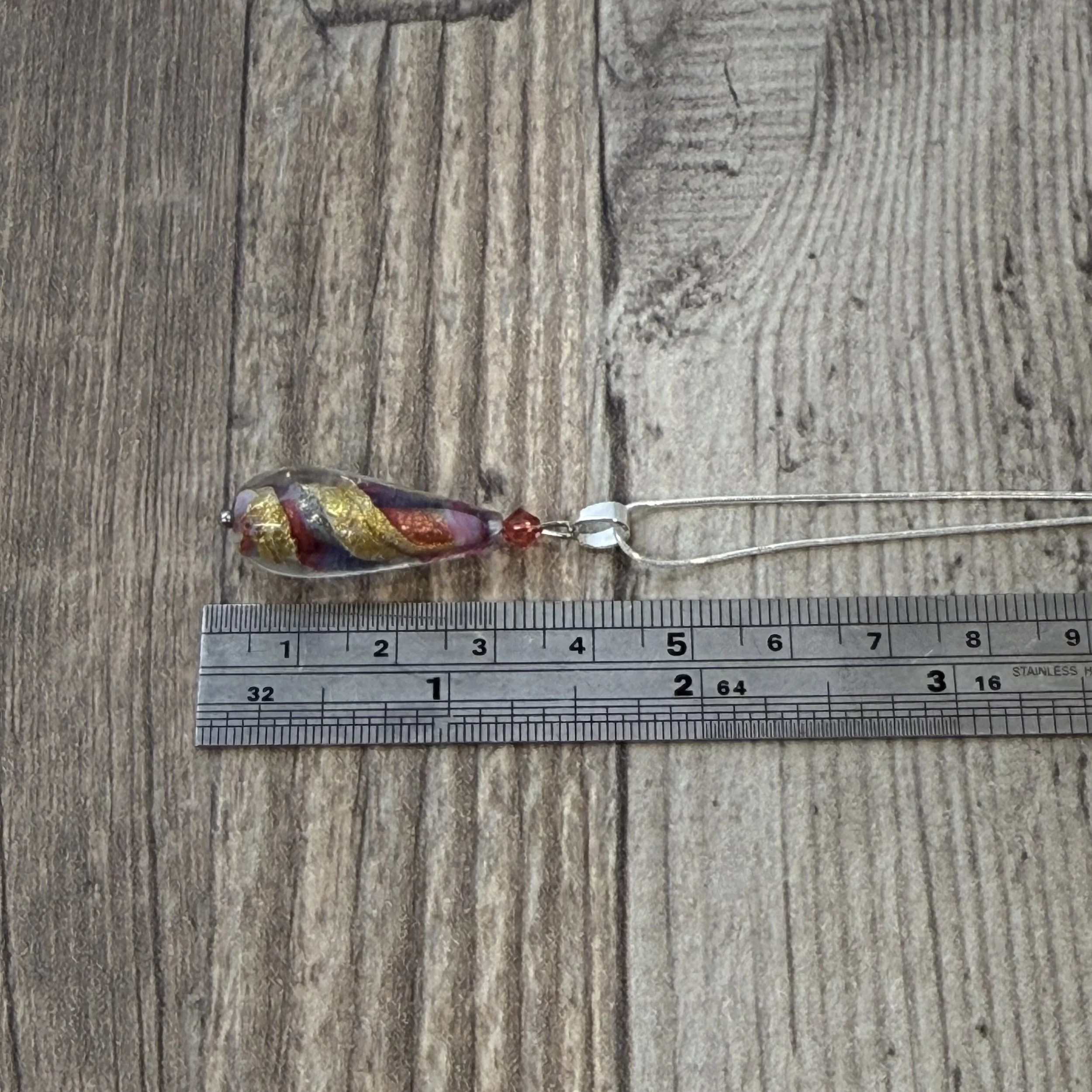 A multi coloured Murano glass teardrop pendant on a sterling silver chain displayed next to a metal ruler on top of a wooden panel