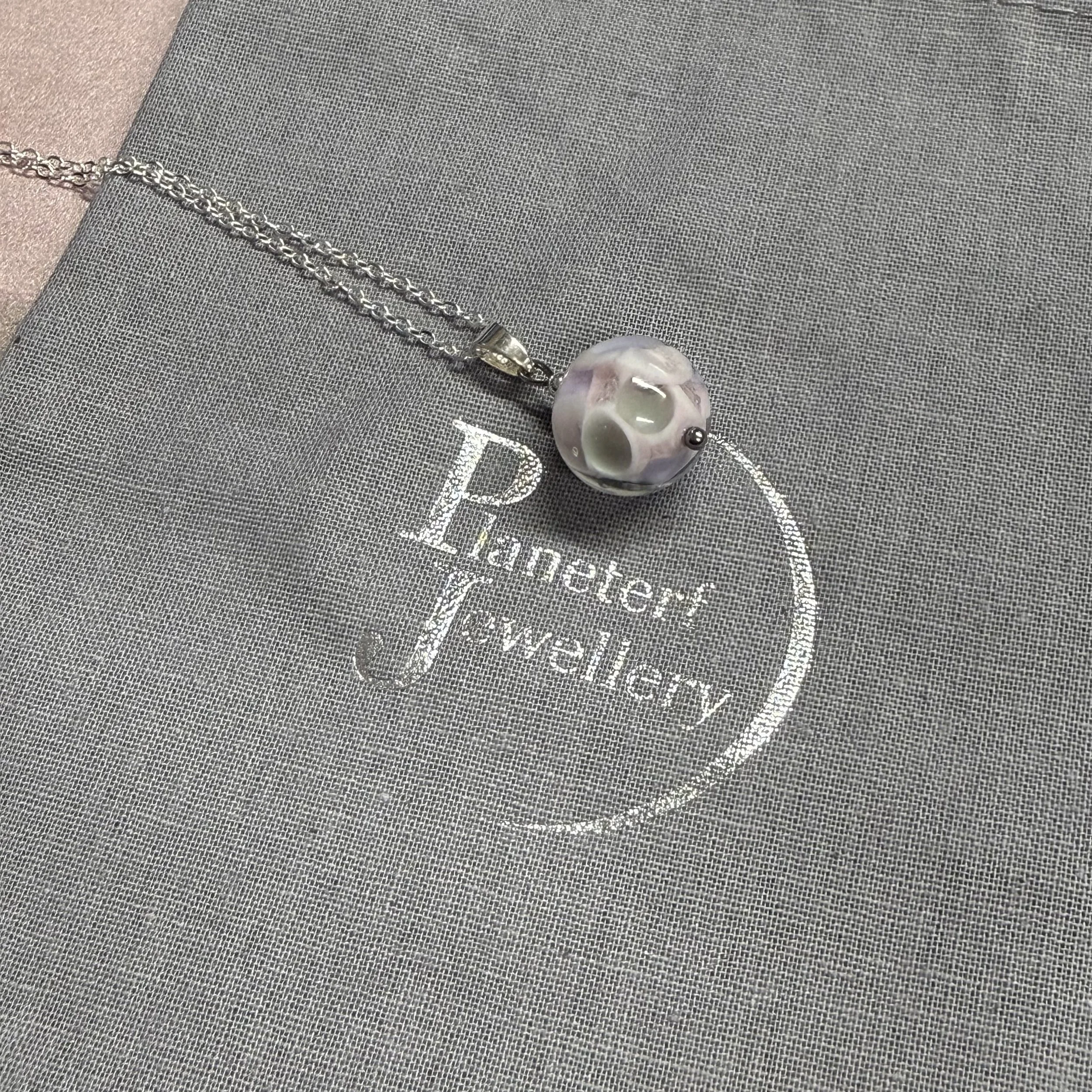 A pink & grey round glass bead hung on a sterling silver chain. The background is a grey cotton bag stamped with Planeterf Jewellery in silver.