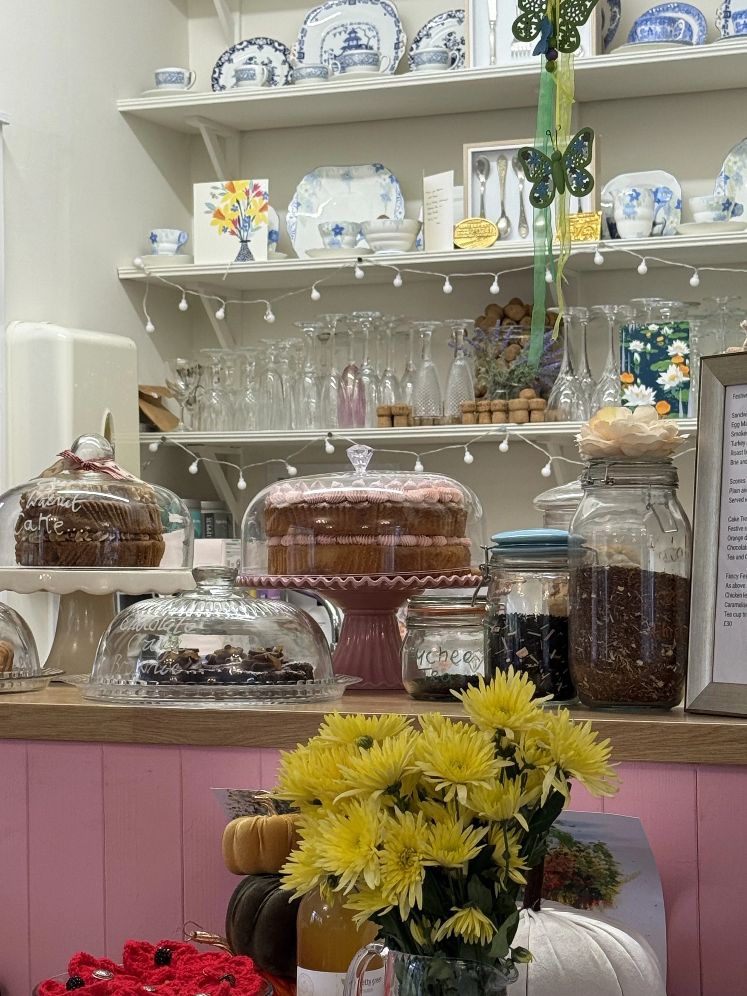 Display of baked goods including cakes, under glass dome covers, on a pink counter with yellow flowers in a vase, set against a background of shelves with blue and white china, glasses, and decorative items.