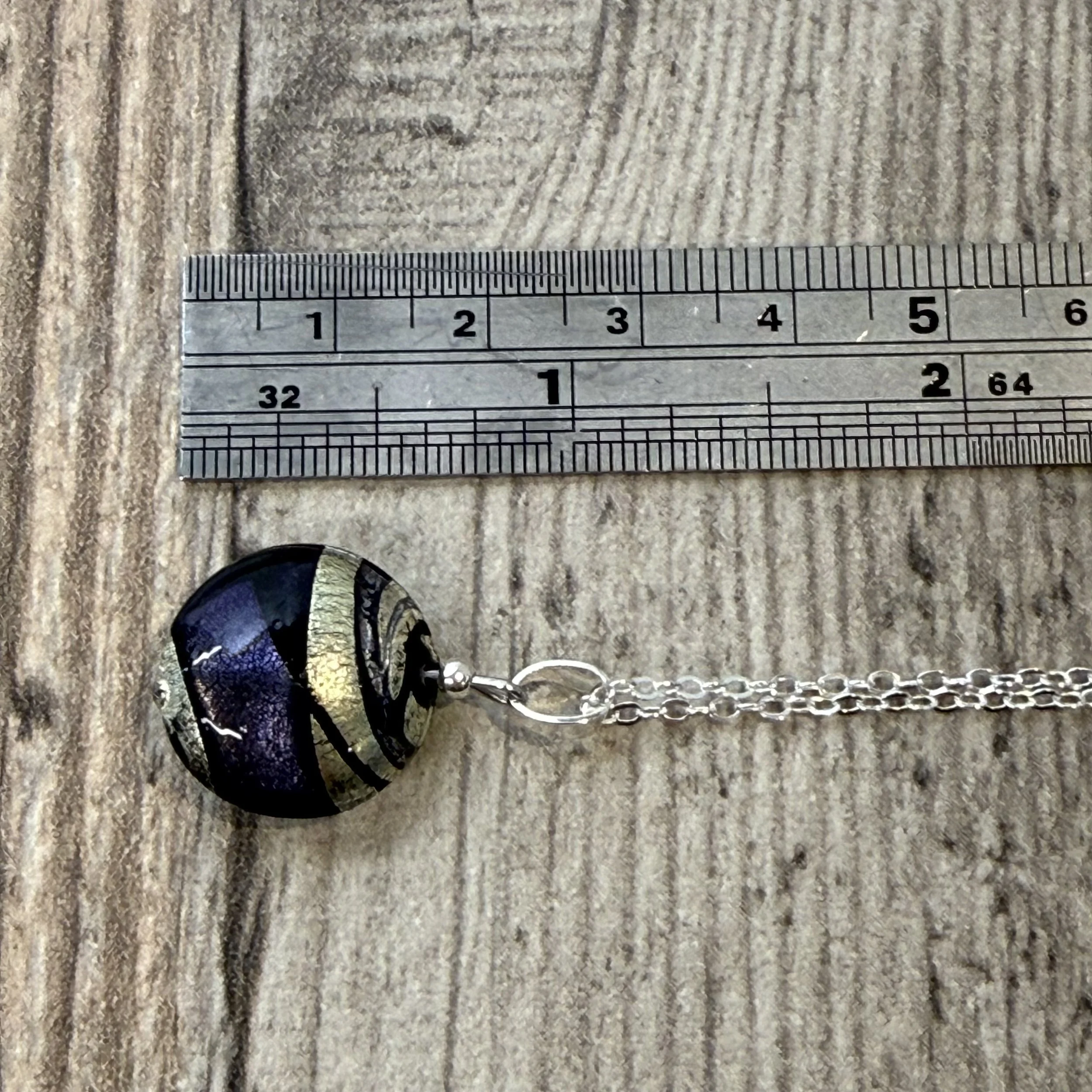 A Murano glass round cushion with purple, black & silver swirls hung on a sterling silver chain next to a metal ruler with a wooden background.