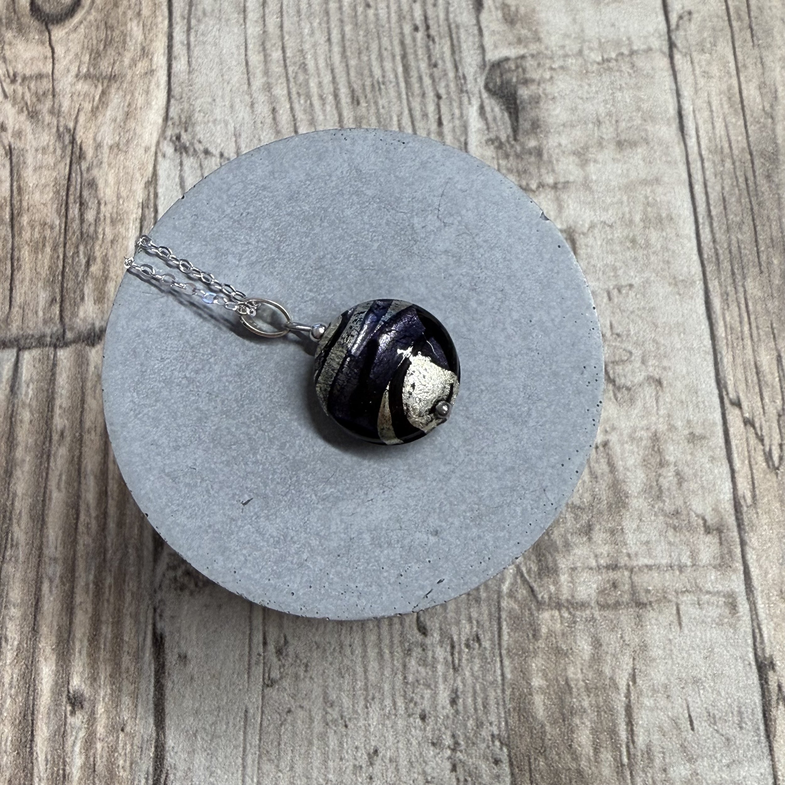 A Murano glass round cushion with purple, black & silver swirls hung on a sterling silver chain. The background is a grey plinth on a wooden base.
