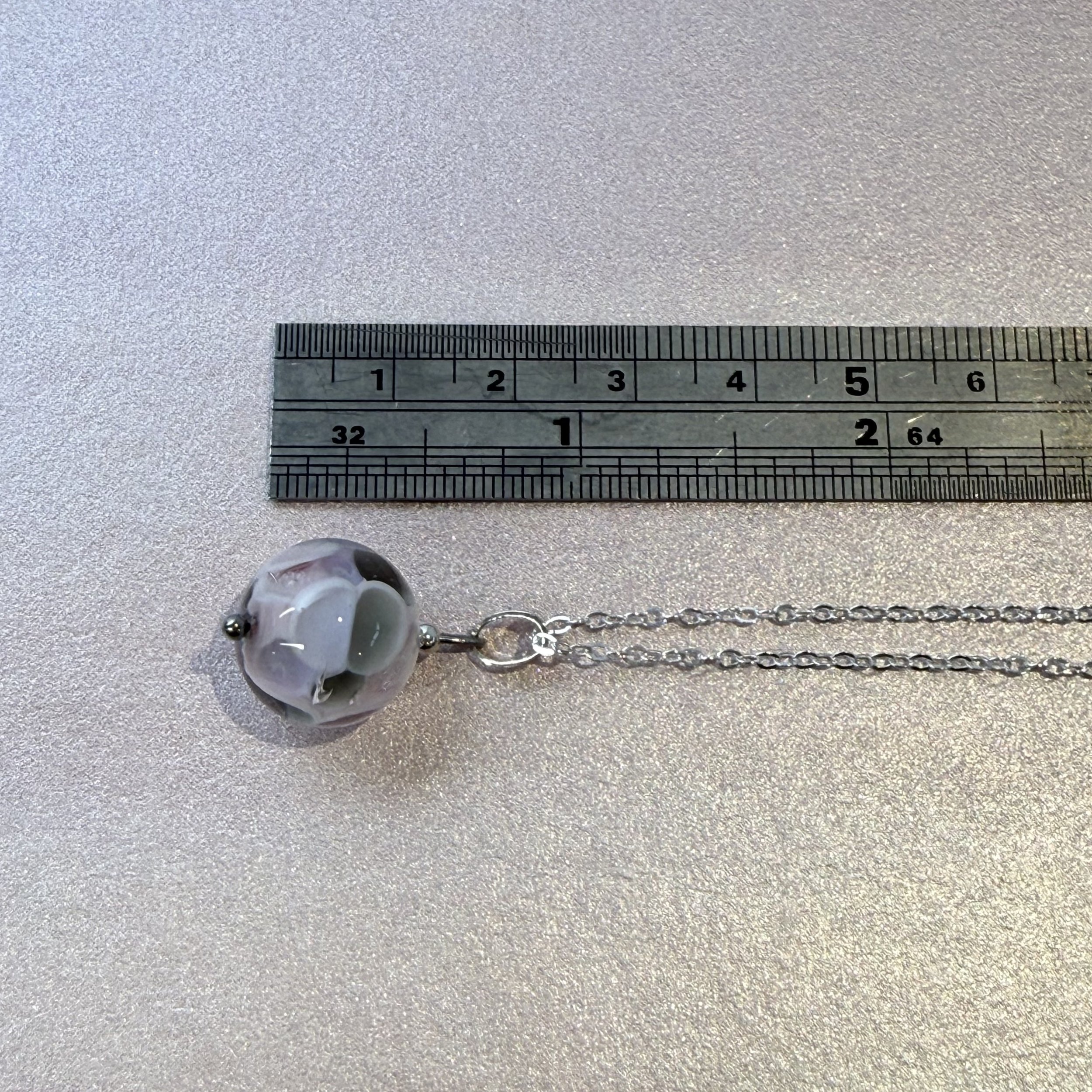 A pink & grey round glass bead hung on a sterling silver chain. next to a metal ruler.