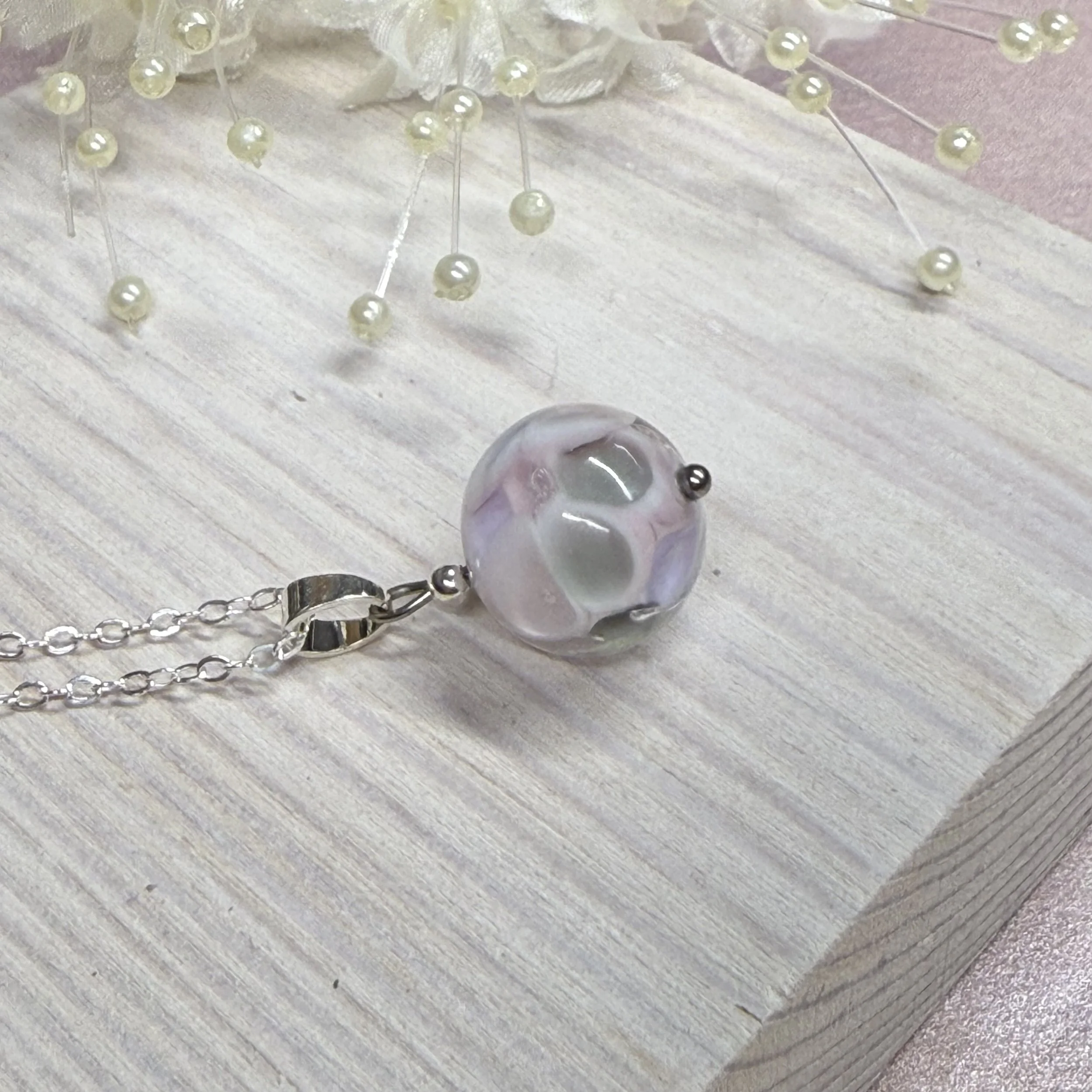 A pink & grey round glass bead hung on a sterling silver chain. The background is a white wooden block with some white floral bits.