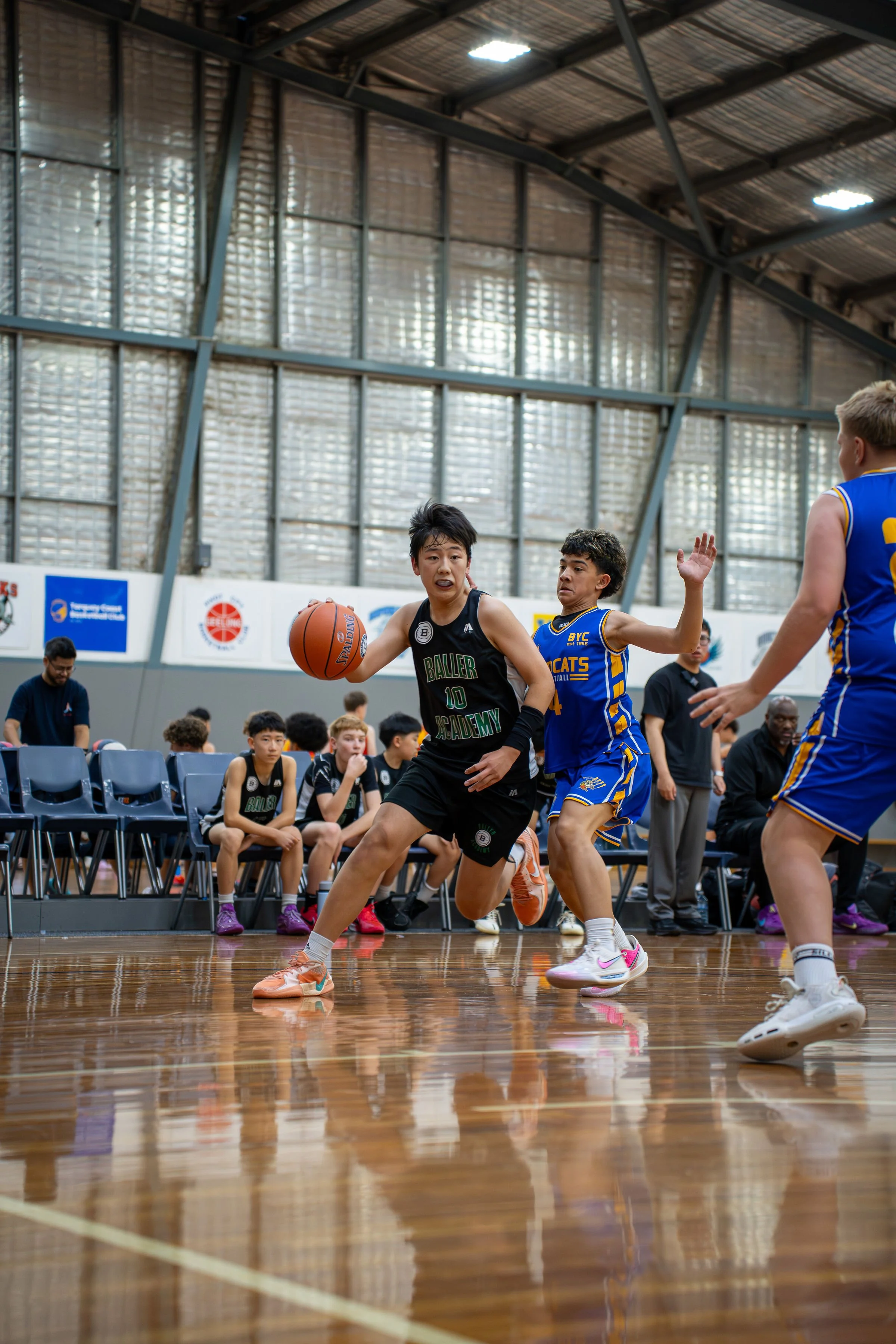 Two young basketball players are competing during a game. One is dribbling the ball, and the other is defending. In the background, several children are seated on chairs, watching the game indoors in a sports facility with a high ceiling and metal walls.