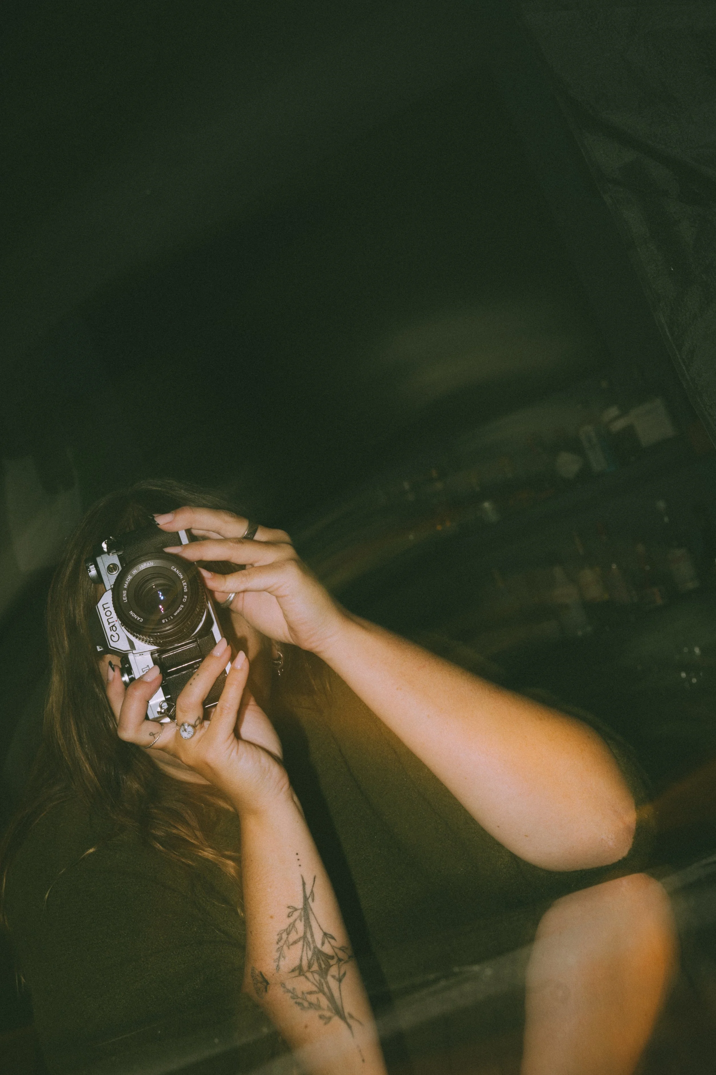 A person with long brown hair, tattoos on the left arm, and nails holding a vintage film camera is taking a photo of themselves.
