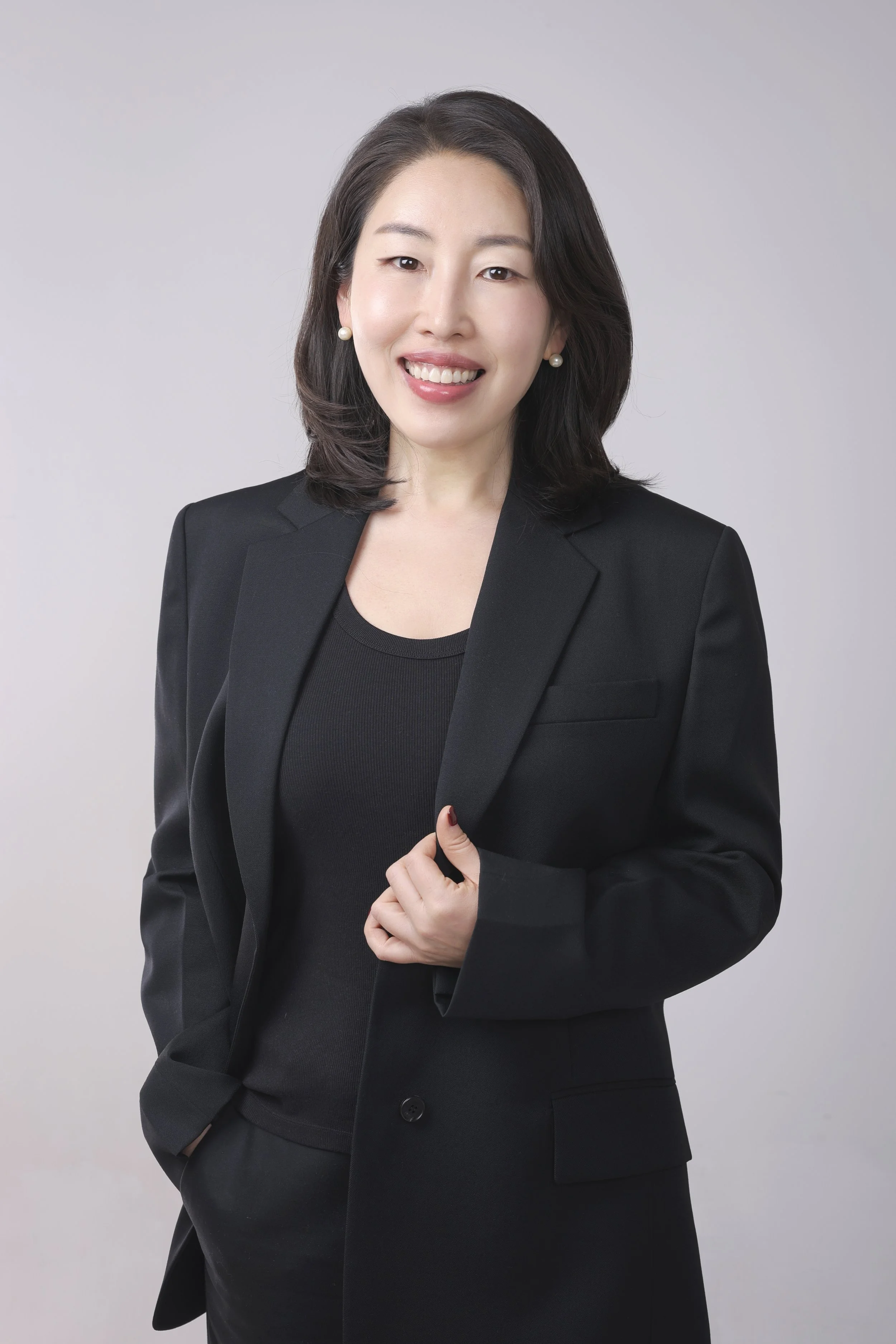 A woman with dark shoulder-length hair, winking, smiling, wearing pearl earrings, a black blazer, and black top, standing against a plain, light grey background.