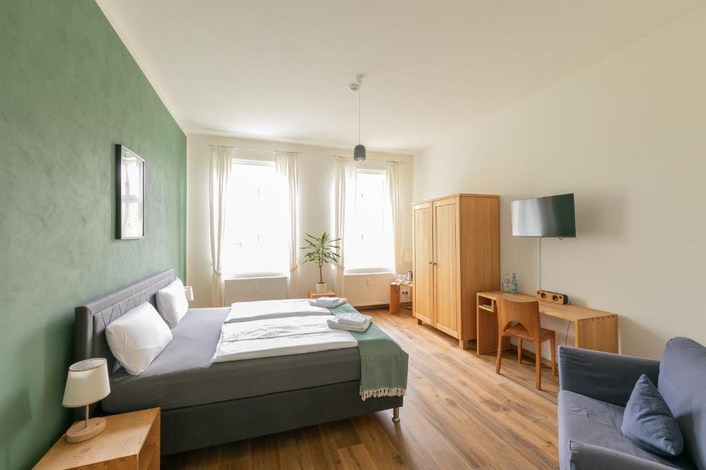 A spacious hotel room with a green accent wall on the left, a large bed with white pillows and linens, a small wooden nightstand and lamp beside the bed, two large windows with white curtains allowing natural light, a potted plant between the windows, a wooden wardrobe, a small desk with a chair, a wall-mounted flat-screen TV, and a gray armchair with cushions on the right.
