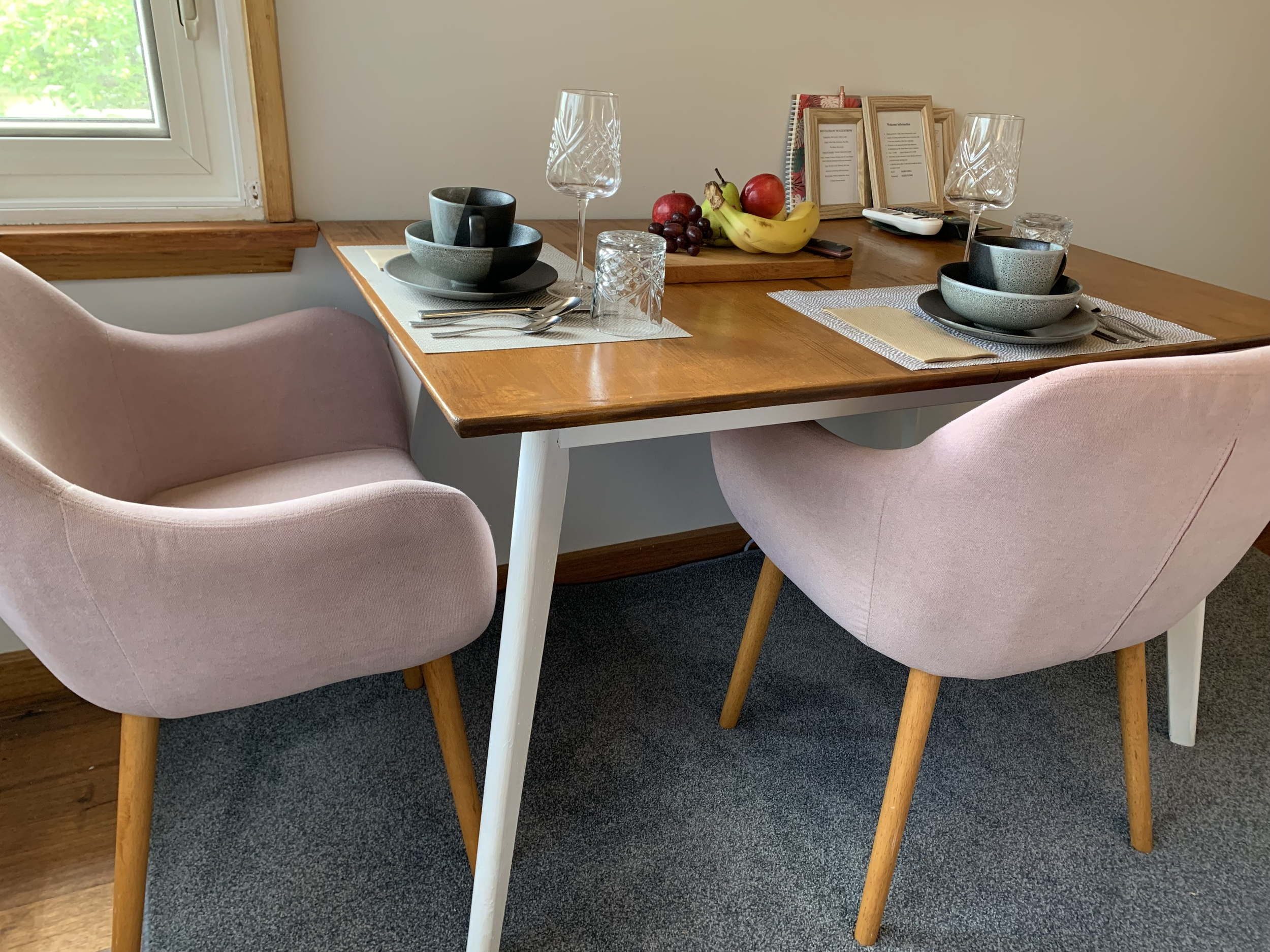 A wooden dining table set for two with pink upholstered chairs, place settings including bowls, glasses, and silverware, a tray of fresh fruit, and framed pictures in the background near a window with greenery outside.