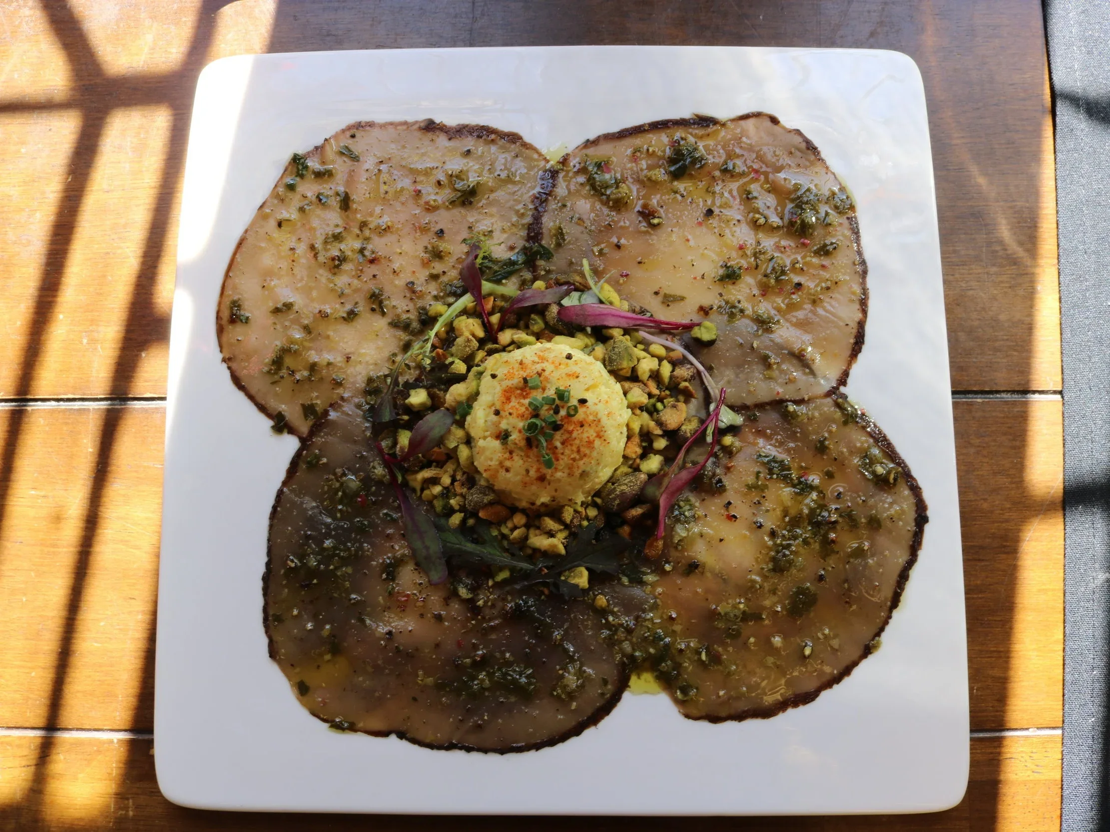 A plate of sliced meat garnished with chopped herbs, crushed pistachios, microgreens, and a dollop of mashed potatoes with paprika, on a wooden table.