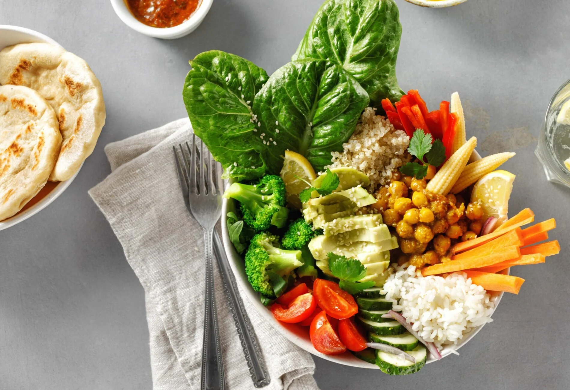 A bowl of colorful vegetable and grain salad with lettuce, cherry tomatoes, cucumber, carrots, chickpeas, rice, avocado, and lemon slices. A small cup of red sauce and a glass of water with lemon are in the background.