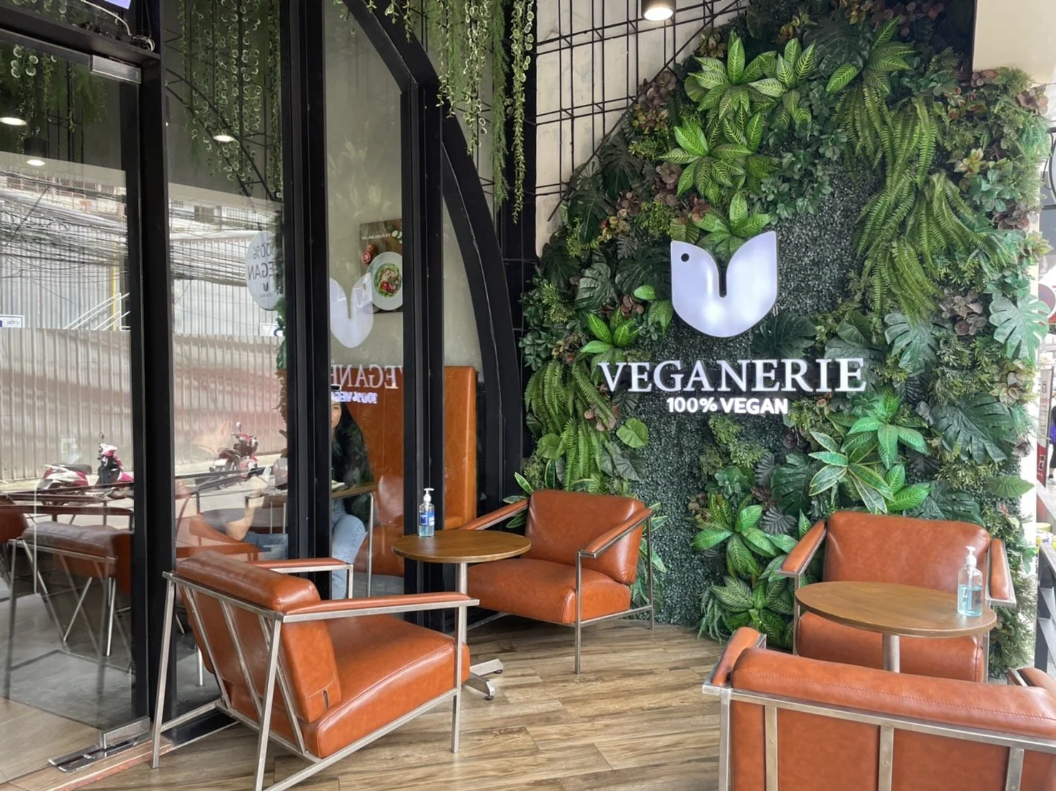 Interior of a vegan restaurant with a green wall, logo, and seating area with orange chairs and small wooden tables.