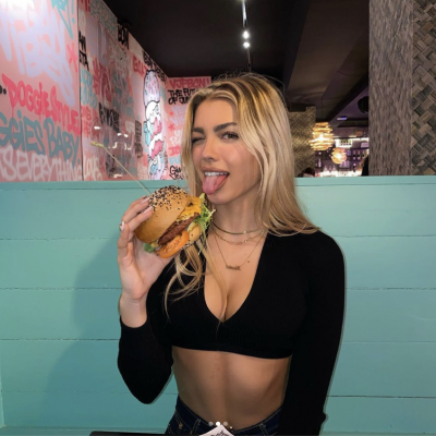 Young woman with blonde hair wearing a black crop top, holding a burger and sticking out her tongue inside a restaurant with colorful graffiti walls.