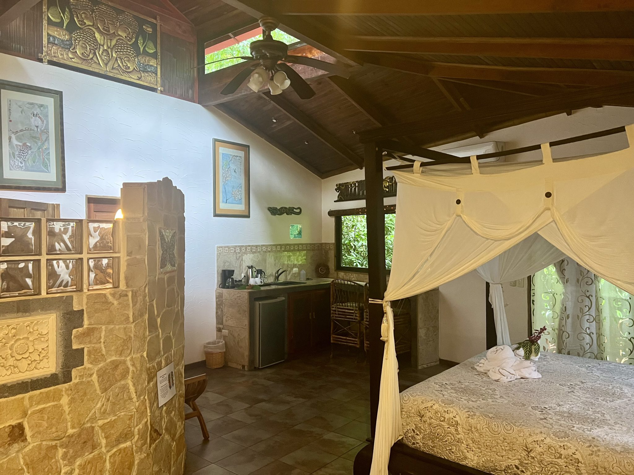 Interior of a tropical-style hotel room with a canopy bed, small kitchenette, stone wall, framed artwork, ceiling fan, and lush green view through the window.