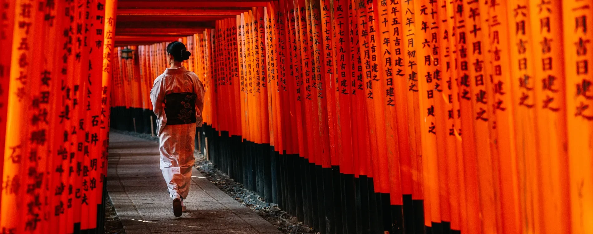A woman dressed in a traditional kimono walking through a tunnel of orange Torii gates at Fushimi Inari Shrine in Japan, with black inscriptions on the gates.