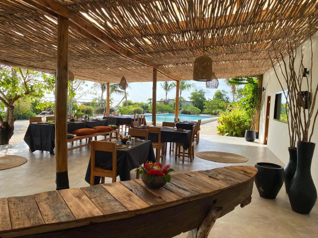 Outdoor dining area with wooden furniture and black tablecloths, overlooking a swimming pool and lush greenery, with a thatched roof and natural decor elements.