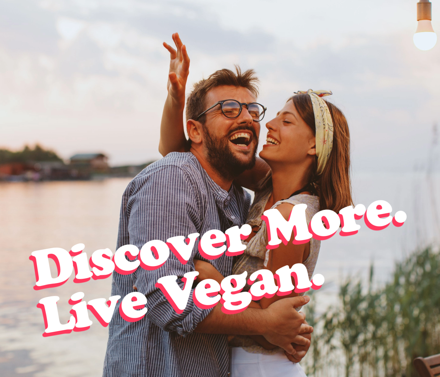 A happy couple hugging near a body of water during sunset, with the words 'Discover More. Live Vegan.' overlaid in bold, pink and white text.