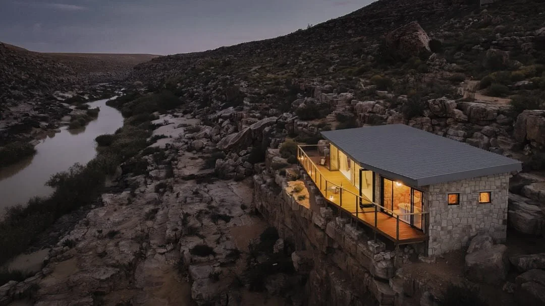 A modern house built on a rocky hillside at dusk, with large windows and a wrap-around balcony overlooking a river and rugged landscape.