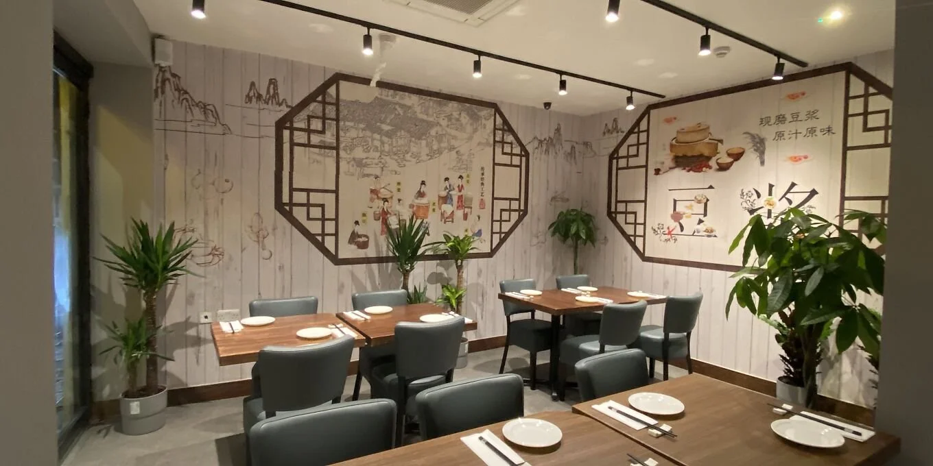 A dining area in a restaurant with wooden tables set with plates and chopsticks. The walls are decorated with traditional Chinese artwork and patterns, and there are tall potted plants around the room.