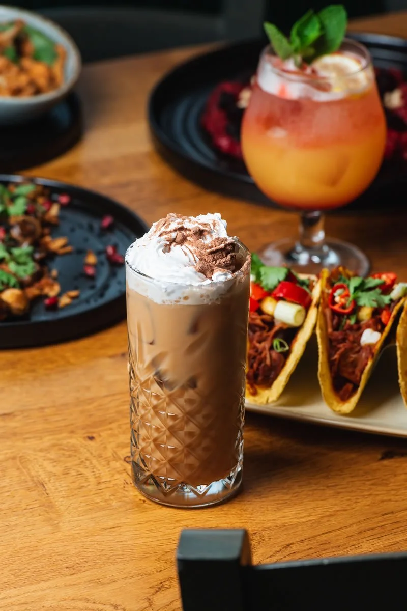 A tall glass of iced coffee topped with whipped cream and chocolate shavings, surrounded by plates of tacos, a bowl of food, and a cocktail with mint garnish on a wooden table.