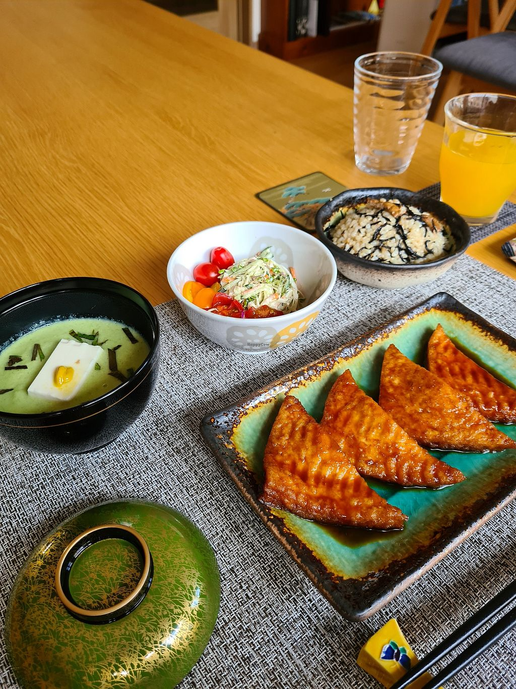 A Japanese meal with fried triangular sushi, a bowl of vinegared rice with seaweed drizzle, a small bowl of chilled cucumber and tomato salad, and a bowl of green miso soup garnished with tofu and seaweed. Two glasses of water and a glass of orange juice are on the table.
