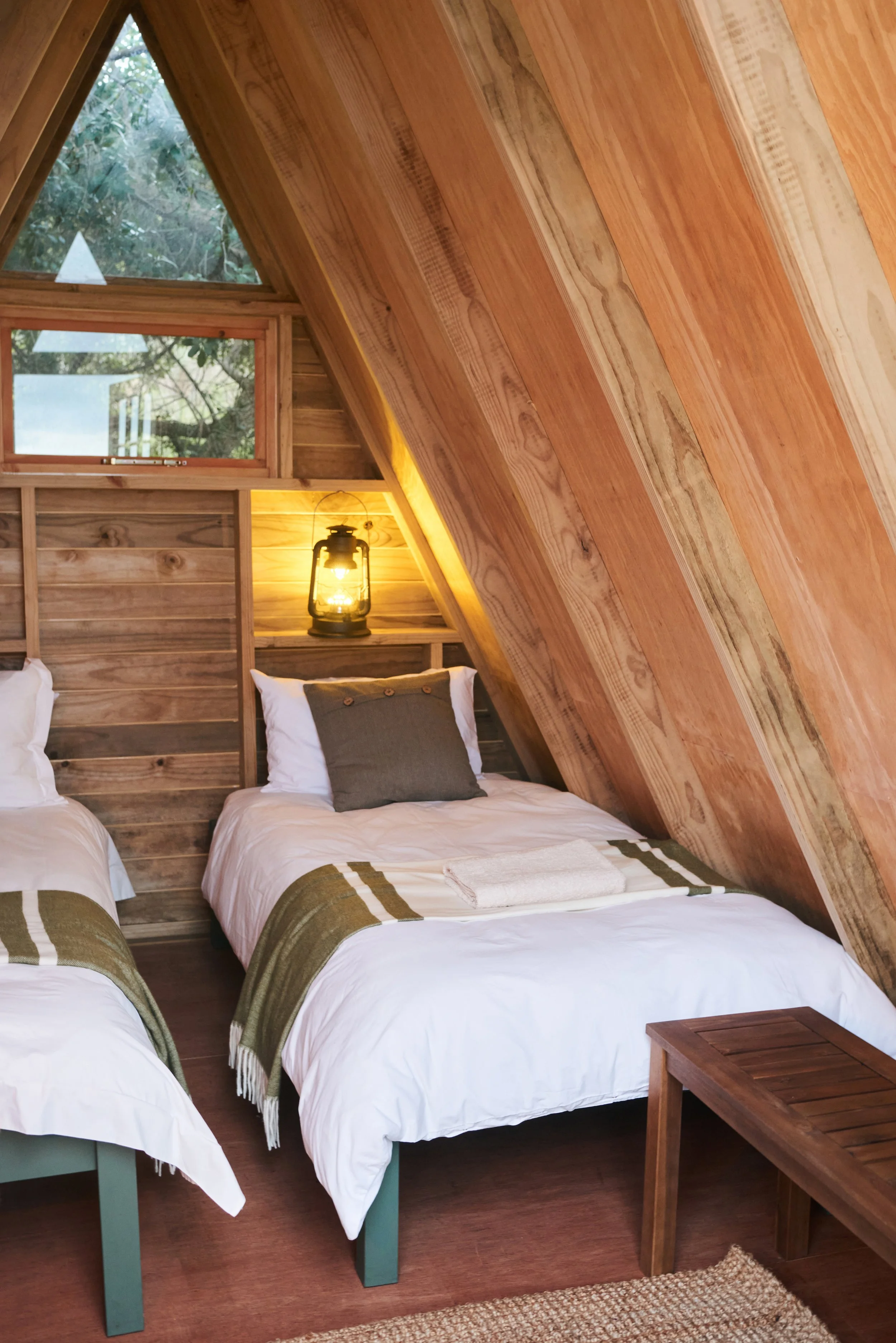 A cozy bedroom with three beds, each with white bedding and green and white blankets, in a room with wooden walls and slanted ceiling. A lantern-style light fixture is mounted on the wall above the beds, and a small wooden bench is positioned to the 