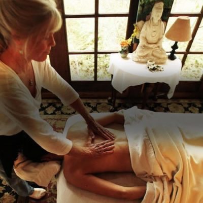 A woman giving a massage to a person lying face down on a massage table in a room with a large window, a statue, and a table with a lamp and flowers.