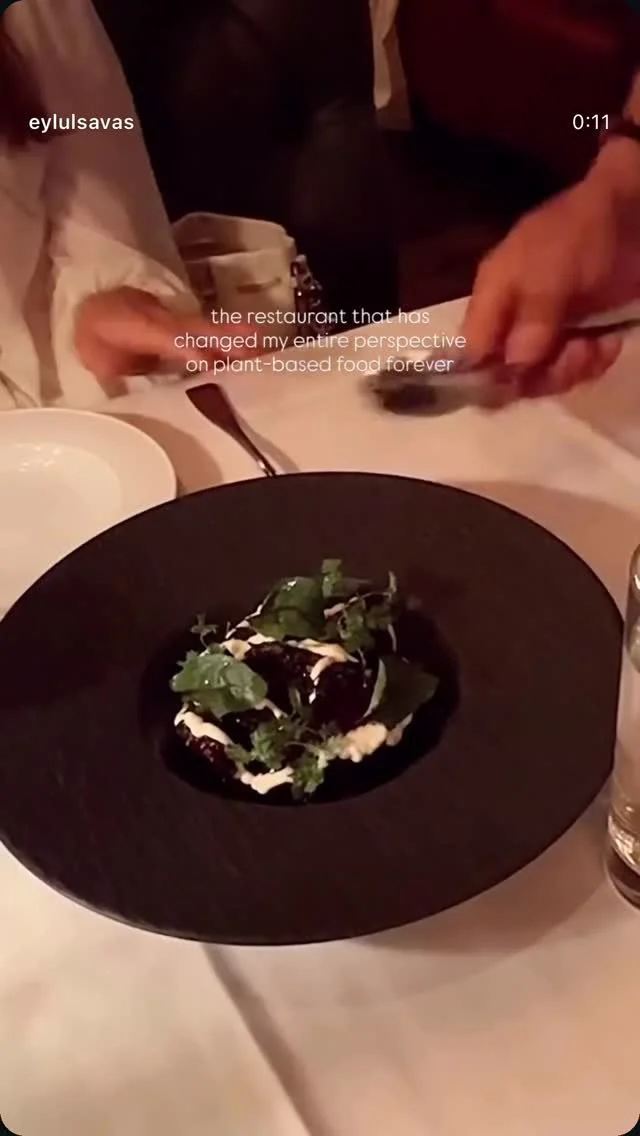 A black plate with a plant-based dish topped with fresh herbs and a white sauce, set on a white tablecloth, with people around the table serving and eating.