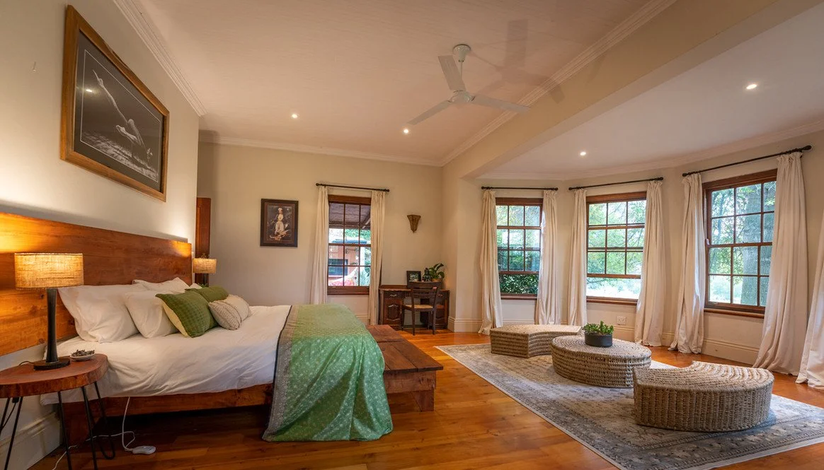 Spacious bedroom with large windows, wooden floors, a bed with green and white bedding, a desk, and cozy wicker chairs with a coffee table.