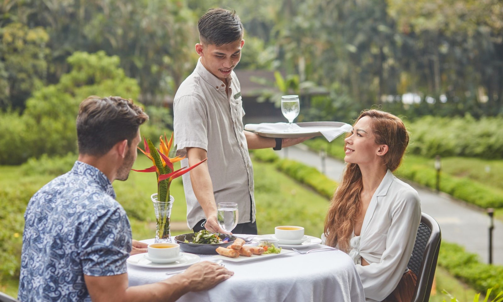 A man in a patterned shirt is sitting at a table with a woman in a white blouse while a waiter serves them a drink outdoors during daytime with greenery in the background.