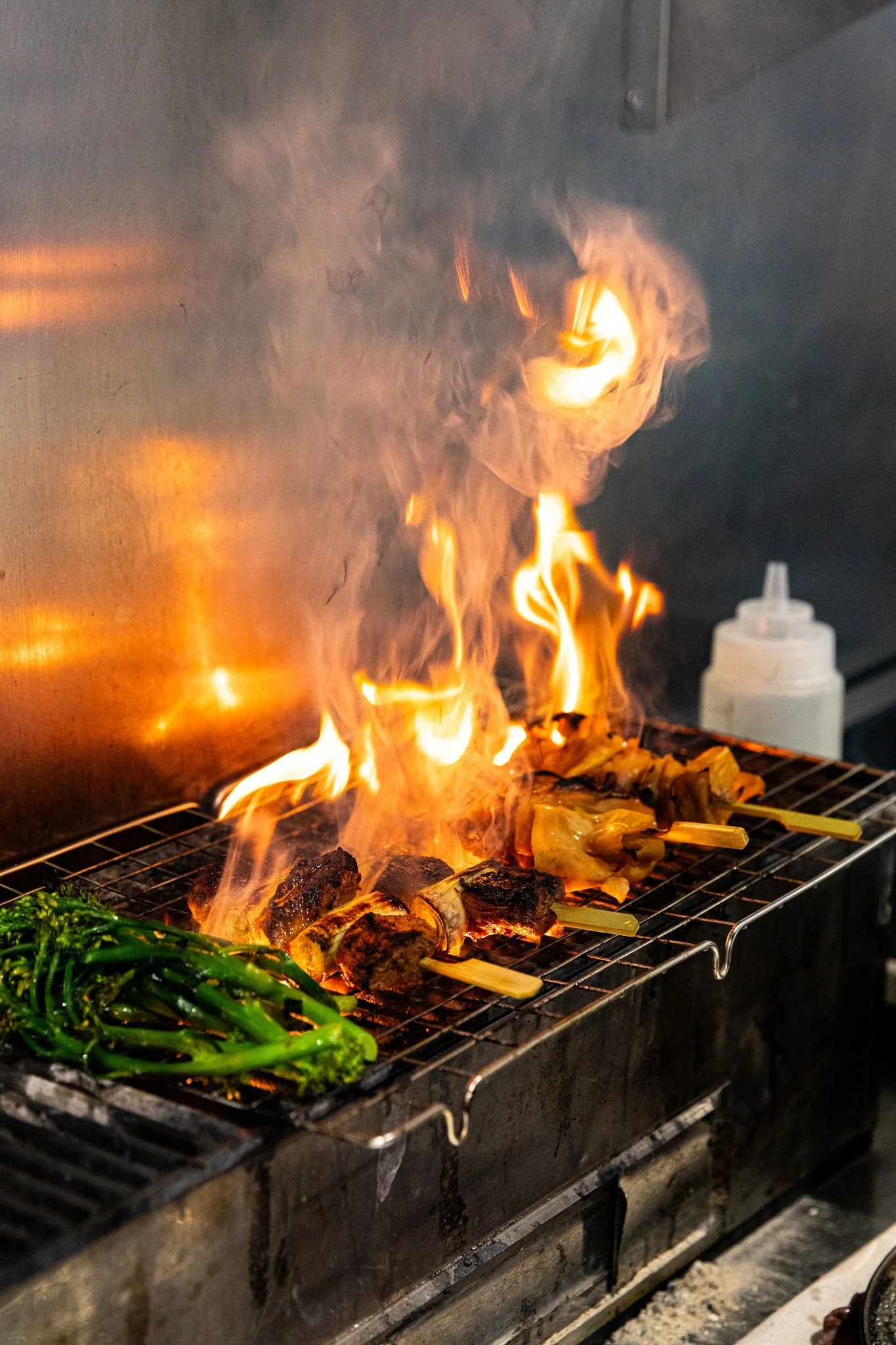 Grill with several skewered meats and green vegetables, some flames and smoke rising from the grill, and nozzles or bottles in the background.