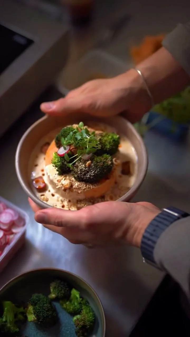 Person holding a bowl of food topped with broccoli, radish, and greens, with other bowls of broccoli and a box of pink circular objects nearby.