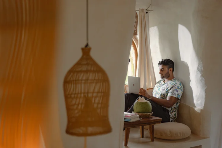 A man sitting on a built-in window seat, working on a laptop, with sunlight coming through a window and nearby decor including a hanging lamp and a stack of books.