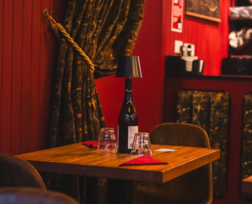 A wooden table in a secluded corner of a restaurant with a wine bottle lamp, two upside-down glasses, and two red napkins, surrounded by dark upholstered chairs and decorative curtains.