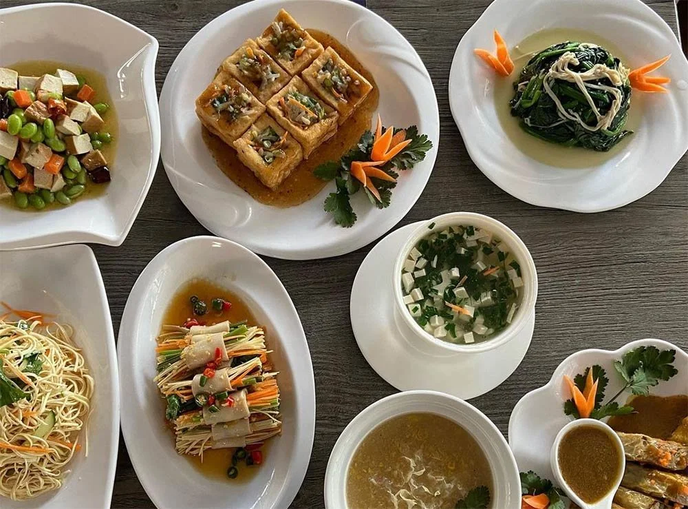 Assorted Chinese dishes on white plates, including stir-fried vegetables, fried tofu with sauce, noodles, vegetable rolls, soup, and garnishes, all arranged on a wooden table.