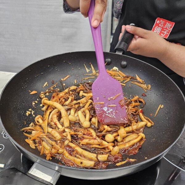 Person cooking stir-fried noodles and vegetables in a black wok using a purple spatula.