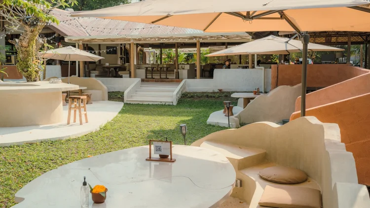 Outdoor patio with white curved seating, umbrellas, and a grassy area in a restaurant or cafe setting.