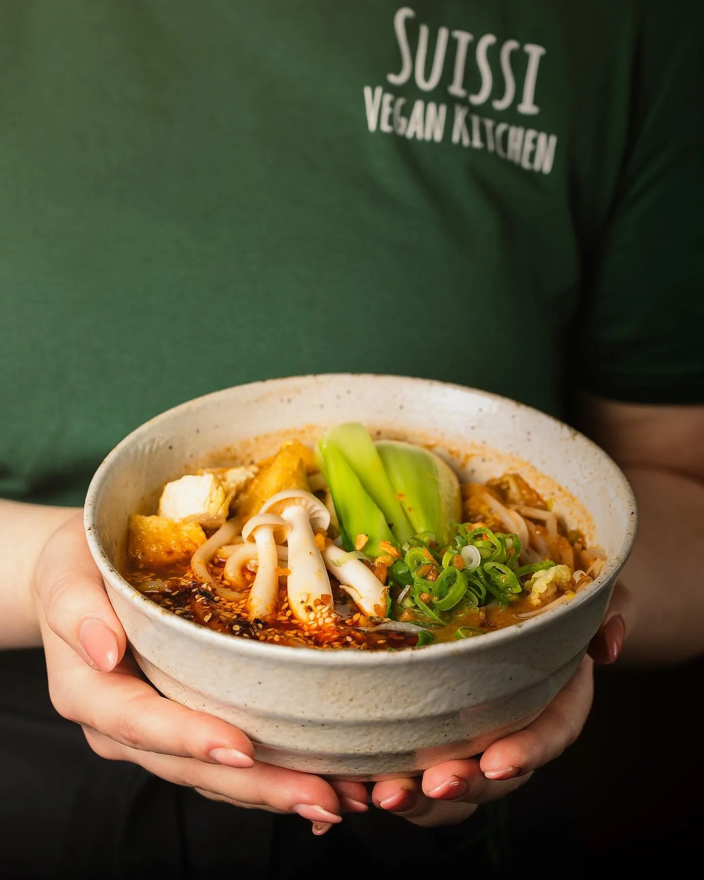 A person holding a bowl of soup with noodles, green vegetables, tofu, and garnishes.